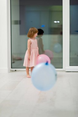 Pembe elbiseli mutlu küçük kız güzel bir akşamda evdeki pencerenin yanında balonlarla oynarken eğleniyor.