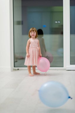 Pembe elbiseli mutlu küçük kız güzel bir akşamda evdeki pencerenin yanında balonlarla oynarken eğleniyor.