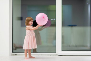 Pembe elbiseli mutlu küçük kız güzel bir akşamda evdeki pencerenin yanında balonlarla oynarken eğleniyor.