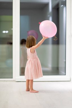 Pembe elbiseli mutlu küçük kız güzel bir akşamda evdeki pencerenin yanında balonlarla oynarken eğleniyor.