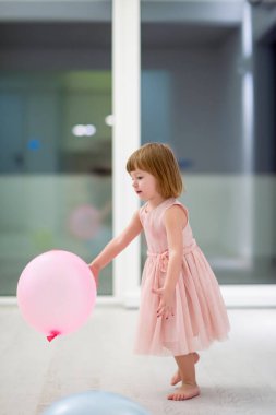 Pembe elbiseli mutlu küçük kız güzel bir akşamda evdeki pencerenin yanında balonlarla oynarken eğleniyor.