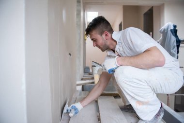 Genç profesyonel inşaat işçisi alçıtaşı duvarlara sıva yapıyor. Yeni, büyük, modern iki katlı apartmanın içinde.