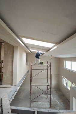Genç profesyonel inşaat işçisi, alçı taşı tavanını sıva ile sıkarken yeni, modern iki katlı apartman dairesinde darağacını kullanıyor.