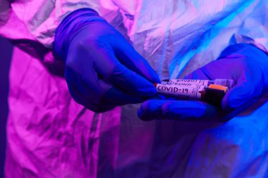 Coronavirus, doktorun elinde kovid 19 virüslü kan örneği test tüpü var. Biyolojik tehlike salgını maskesi, giysisi ve parlak ışık arkaplanı var.
