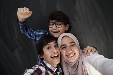 Bir grup Arap genci arka planda siyah karatahta ile akıllı telefonlarda selfie çekiyorlar.
