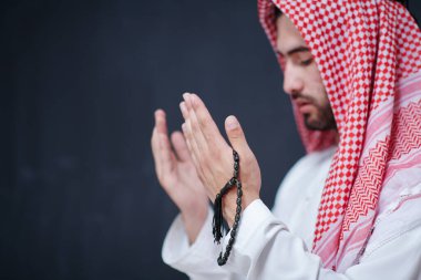 Geleneksel dua eden geleneksel giysili genç Arap adam, modern İslami modayı ve Ramazan Kareem kavramını temsil eden siyah karatahtanın önünde el ele dua ediyor.