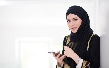 Genç, modern Müslüman iş kadını akıllı telefon kullanıyor geleneksel tesettür kıyafetlerini evde giyiyor.