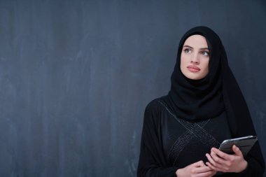 Genç, modern Müslüman iş kadını elinde tablet bilgisayarla siyah karatahta önünde tesettürlü kıyafetler giyiyor.