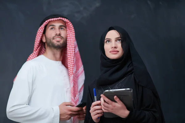 若いイスラム教徒のビジネスカップルアラビア人男性と女性でファッショナブルなヒジャーブのドレス携帯電話やタブレットコンピュータを使用して黒の黒板の前に現代のイスラムファッション技術とラマダーンKareem概念を表す — ストック写真