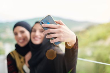 Modern İslami moda teknolojisi ve Ramazan kareem kavramını temsil eden balkonda selfie çekerken, tesettürlü şık giyinmiş genç, güzel Müslüman kadınlar cep telefonu kullanıyor.