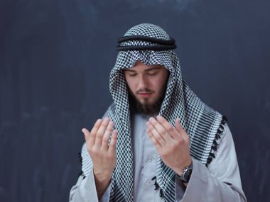 Geleneksel dua eden geleneksel giysili genç Arap adam, modern İslami modayı ve Ramazan Kareem kavramını temsil eden siyah karatahtanın önünde el ele dua ediyor.