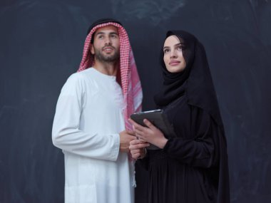 Modern İslami moda teknolojisi ve ramazan kareem kavramını temsil eden siyah karatahtanın önünde cep telefonu ve tablet bilgisayar kullanan, modaya uygun tesettürlü bir kadınla genç Müslüman çift.
