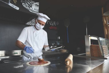 Modern restoran mutfağında özel ahşap fırınıyla geleneksel İtalyan pizzası hazırlayan yetenekli bir şef. Korunaklı tıbbi yüz maskesi ve koronavirüs eldivenleri takmak. Yeni normal kavram.