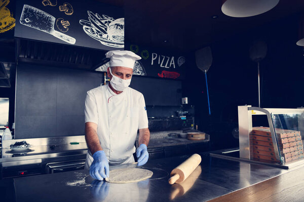 Skilled chef preparing traditional italian pizza in interior of modern restaurant kitchen with special wood-fired oven. Wearing protective medical face mask and gloves in coronavirus new normal concept