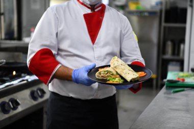 Şef aşçı, Corona virüsü hastalığından korunmak için yüz koruyucu maske takıyor ve profesyonel restoran mutfağında bir tabak otantik sandviç yiyor. Yeni normal konseptte sağlık, güvenlik ve salgın konsepti