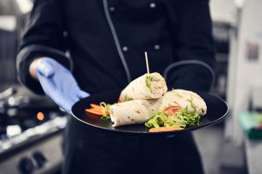 Şef aşçı, Corona virüsü hastalığından korunmak için yüz koruyucu maske takıyor ve profesyonel restoran mutfağında bir tabak otantik sandviç yiyor. Yeni normal konseptte sağlık, güvenlik ve salgın konsepti
