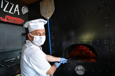 Modern restoran mutfağında özel ahşap fırınıyla geleneksel İtalyan pizzası hazırlayan yetenekli bir şef. Korunaklı tıbbi yüz maskesi ve koronavirüs eldivenleri takmak. Yeni normal kavram.
