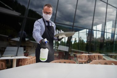 Garson, restoranda dezenfektan spreyiyle masayı temizliyor koruyucu tıbbi maske ve eldiven takıyor.