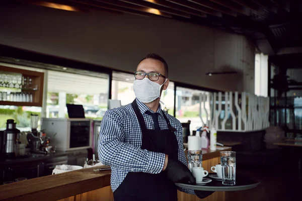 waiter in a medical protective mask serves  the coffee in restaurant durin coronavirus pandemic representing new normal concept