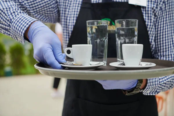 waiter in a medical protective mask serves  the coffee in restaurant durin coronavirus pandemic representing new normal concept