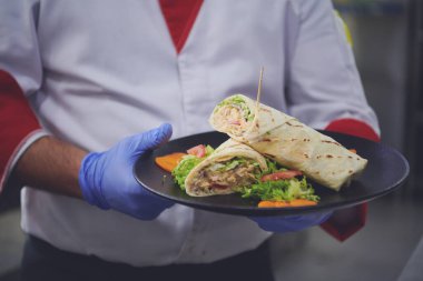 Şef aşçı, Corona virüsü hastalığından korunmak için yüz koruyucu maske takıyor ve profesyonel restoran mutfağında bir tabak otantik sandviç yiyor. Yeni normal konseptte sağlık, güvenlik ve salgın konsepti