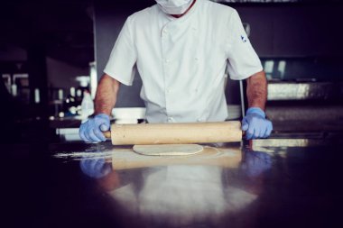Modern restoran mutfağında özel ahşap fırınıyla geleneksel İtalyan pizzası hazırlayan yetenekli bir şef. Korunaklı tıbbi yüz maskesi ve koronavirüs eldivenleri takmak. Yeni normal kavram.