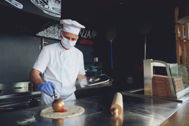 Modern restoran mutfağında özel ahşap fırınıyla geleneksel İtalyan pizzası hazırlayan yetenekli bir şef. Korunaklı tıbbi yüz maskesi ve koronavirüs eldivenleri takmak. Yeni normal kavram.