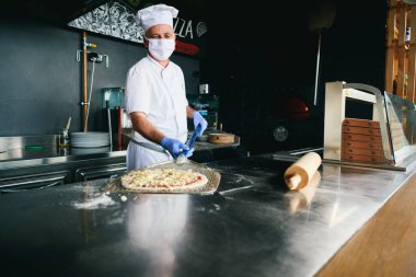 Modern restoran mutfağında özel ahşap fırınıyla geleneksel İtalyan pizzası hazırlayan yetenekli bir şef. Korunaklı tıbbi yüz maskesi ve koronavirüs eldivenleri takmak. Yeni normal kavram.