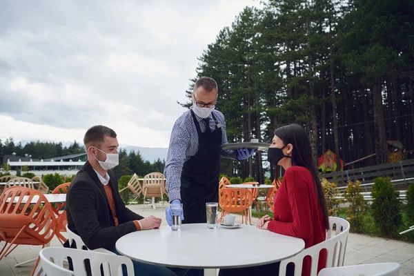 Waiter with protective medical mask and gloves serving guest with coffee at an outdoor bar cafe or restaurant new normal concept reopening after quarantine