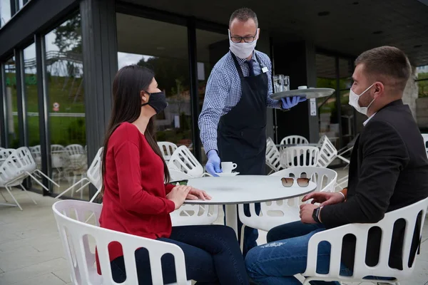 Waiter with protective medical mask and gloves serving guest with coffee at an outdoor bar cafe or restaurant new normal concept reopening after quarantine