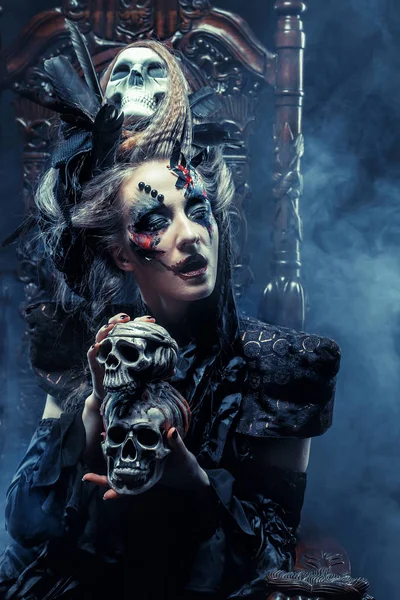 Young beautiful witch sits on a chair. Bright make up, skull, smoke-  halloween theme. — Stock Photo, Image