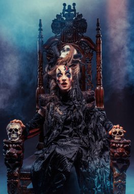 Genç ve güzel cadı bir sandalyeye oturur. Parlak makyaj, kafatası, sigara... Cadılar Bayramı teması.