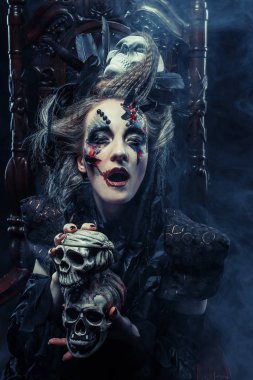 Genç ve güzel cadı bir sandalyeye oturur. Parlak makyaj, kafatası, sigara... Cadılar Bayramı teması.