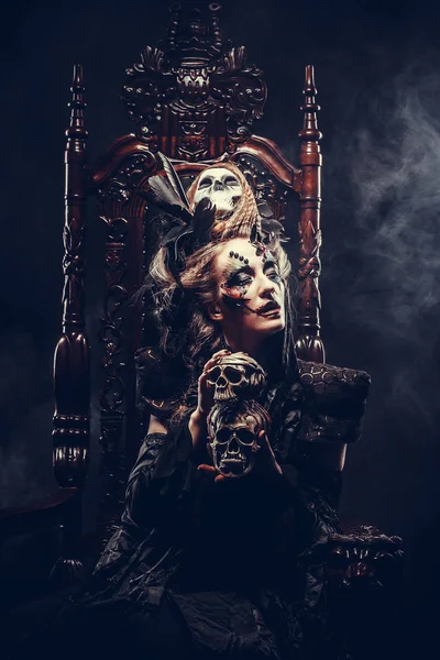 Mladá krásná čarodějka sedí na židli. Jasné make-up, lebka, kouř- halloween téma. — Stock fotografie