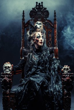 Genç ve güzel cadı bir sandalyeye oturur. Parlak makyaj, kafatası, sigara... Cadılar Bayramı teması.