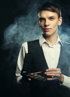 Genç erkek makyaj sanatçısı elinde gölge paleti ve fırça tutuyor..