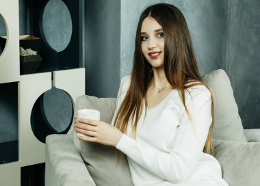 Güzel ve sevimli. Elinde kahve fincanı tutan ve evdeki sandalyeye oturan güzel genç bir kadın. Yaşam tarzı kavramı.