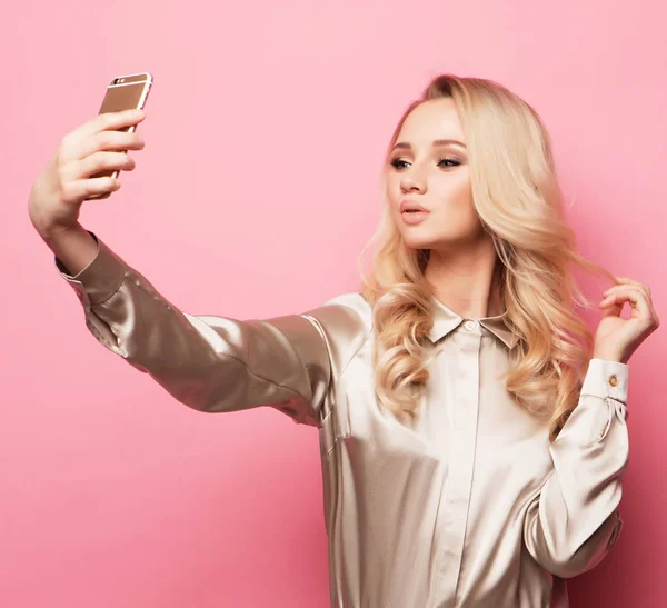 Портрет красивой улыбающейся блондинки в повседневной одежде с идеальным макияжем со смартфоном, делает селфи на розовом фоне — стоковое фото