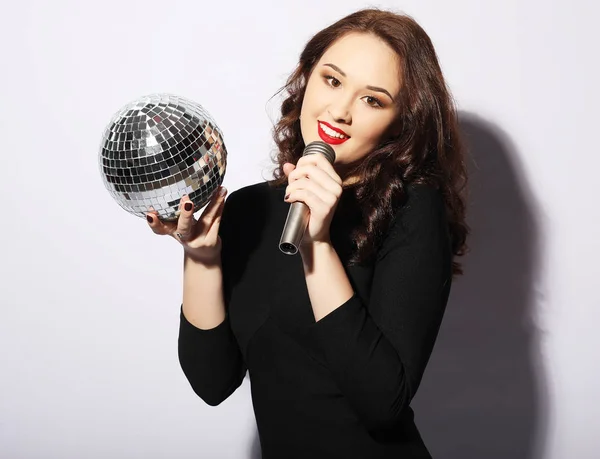 Jovem mulher vestindo vestido preto, segurando bola de discoteca e cantando no microfone — Fotografia de Stock