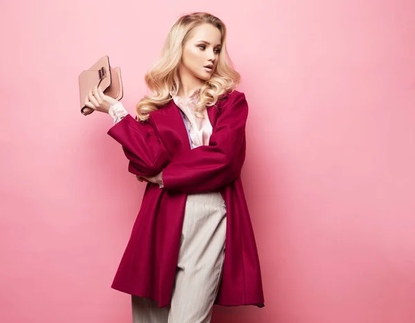 Mulher loira bonita em uma blusa e calças usando óculos, segurando bolsa posando sobre fundo rosa. — Fotografia de Stock