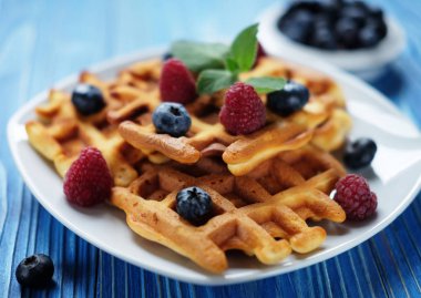 Geleneksel Belçika waffleları taze böğürtlenler ve beyaz tabakta şeker tozu, mavi ahşap arka plan. Düz konum, üst görünüm, kopyalama alanı.