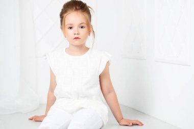 Çocuk moda. Oturma ve poz Beyaz giysili güzel dalgın ciddi sarışın kızı.