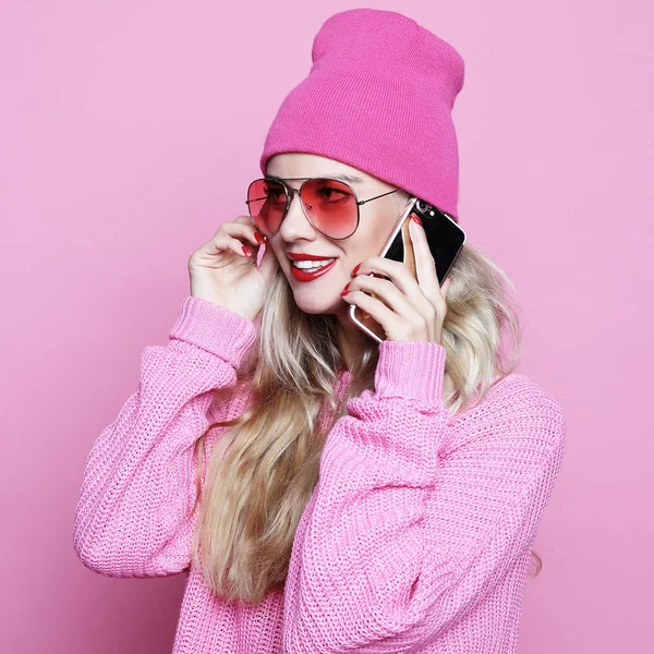 时尚快乐凉爽的微笑的女孩谈论在粉红色的衣服在粉红色的背景下, 穿着粉红色的帽子粉红色的太阳镜和粉红色的羊毛衫智能手机. — 图库照片