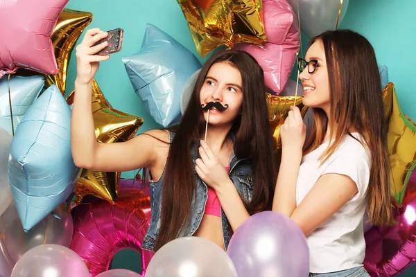 Två tonårstjejer med färgglada ballonger gör selfie på en telefon. Blå bakgrund. — Stockfoto