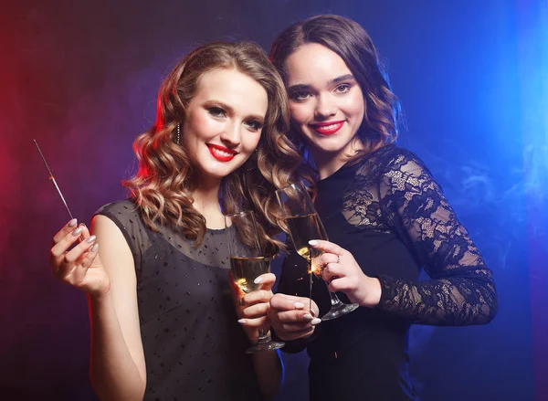 Deux belles jeunes femmes en robe noire avec verres à vin et feu scintillant . — Photo
