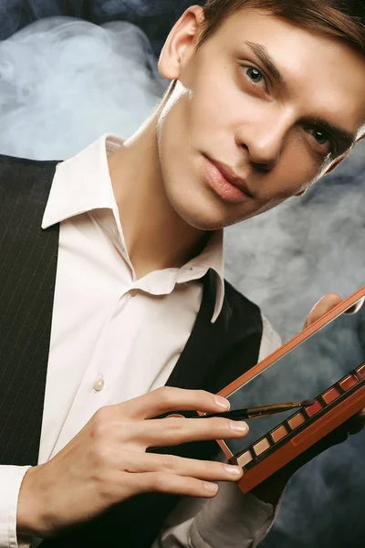 Professional makeup artist posing in studio, holds a palette of shadows. Against the background of smoke.