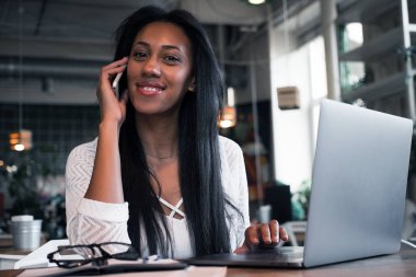 yaşam tarzı ve insanlar kavramı: cep telefonu ve dizüstü bilgisayar kullanarak bir kafede mutlu genç Afrikalı kadın