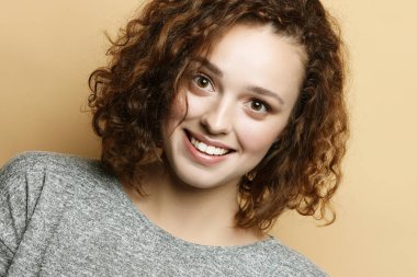 Güzel, kıvırcık, tatlı gülüşlü bir kadın bej arka planda yalnız poz veriyor..