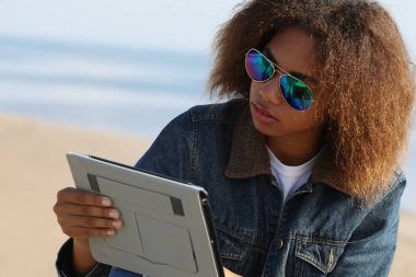 Plajda tableti olan Afrikalı Amerikalı genç bir kadın.