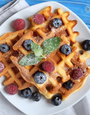 Geleneksel Belçika waffleları taze böğürtlenler ve beyaz tabakta şeker tozu, mavi ahşap arka plan. Düz konum, üst görünüm, kopyalama alanı.